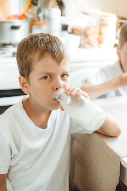 男の子が自宅のキッチンでボトルからミルクを飲んでいます。ミルクと朝の朝食。白いTシャツを着た幸せな子供はボトルからミルクを飲みます