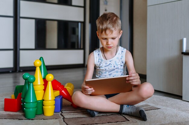 The boy is in the game room, next to toys, but does not play, but looks at the tablet