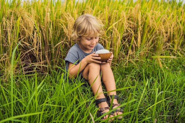 Boy is holding a cup of boiled rice in a wooden cup on the background of a ripe rice field. Food for children concept.