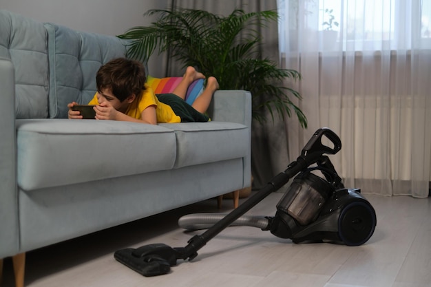 Boy is lying on the couch and playing on the phone next to him on the floor is a vacuum cleaner