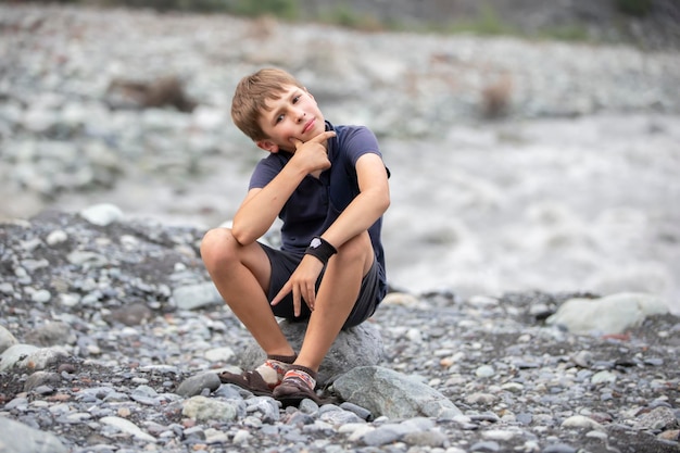 The boy is sitting on the bank of a mountain river