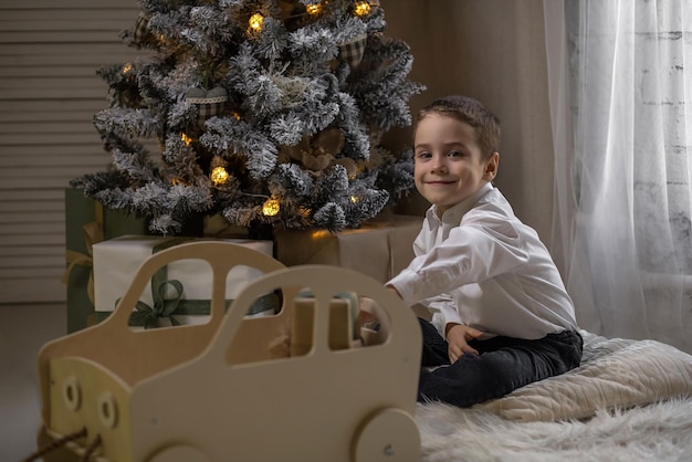 Foto il ragazzo è seduto sul tappeto vicino all'albero di natale con in mano un regalo e gioca con una macchina da scrivere in legno