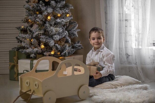 Foto il ragazzo è seduto sul tappeto vicino all'albero di natale con in mano un regalo e gioca con una macchina da scrivere in legno
