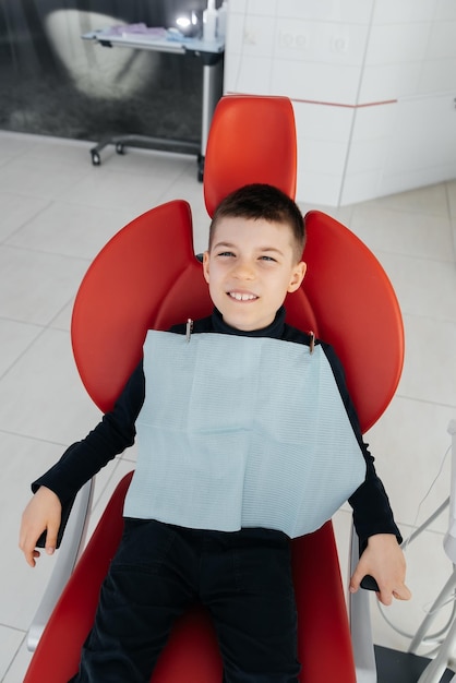 The boy is sitting in a red dental chair and smiling in modern\
white dentistry treatment and prevention of caries since childhood\
modern dentistry and prosthetics