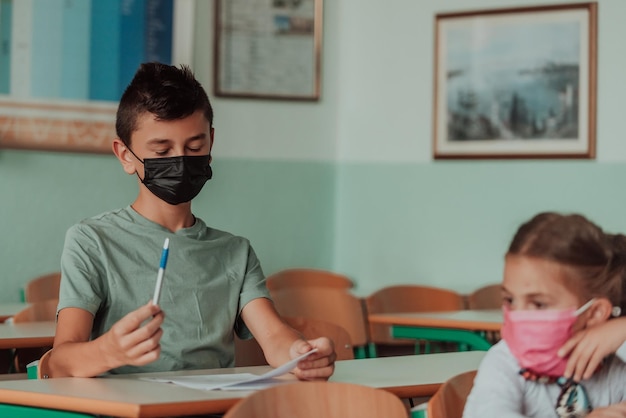 그 소년은 학교 책상에 앉아 코로나 바이러스 보호에 맞서 얼굴에 마스크를 쓰고 있습니다.