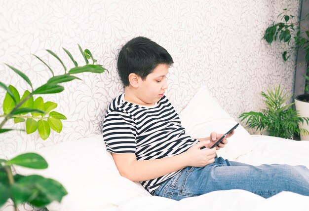 A boy is watching a digital tablet and smiling on light bedroom under blanket