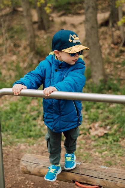 A boy in a jacket and sunglasses stands in the park leaning on the crossbar