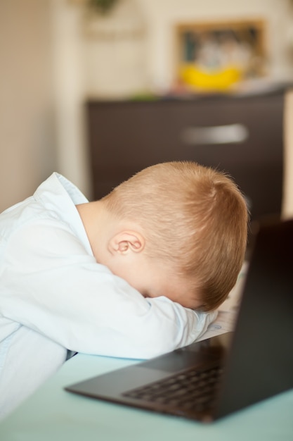 デジタルタブレットのラップトップノートブックを机の上に座っている男の子の子供。彼はとても疲れて眠りについた。子供のオンライン学習。オンライン教育の遠隔学習。