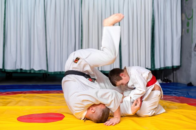 A boy in kimono is practicing martial arts with his master in the ring of a gym, boy is winning