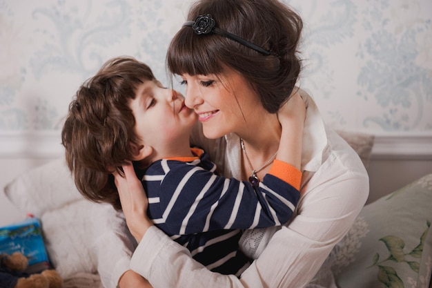 Мальчик целует свою маму и обнимает. Счастливая молодая женщина весело провести время с мальчиком милый ребенок. Мама и сын сидят на кровати перед сном. Концепция спокойной ночи