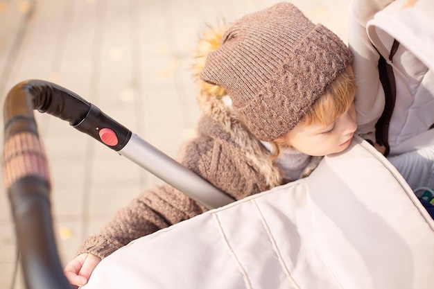 a boy in a knitted hat and a brown jacket looks at a baby stroller on the street