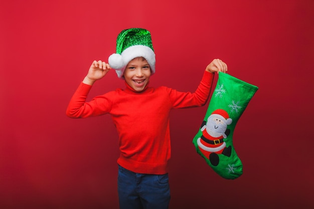 A boy in a knitted red sweater and a Santa Claus hat holds a Christmas boot for gifts Cute baby is waiting for Christmas and a new kind insulation on a red background