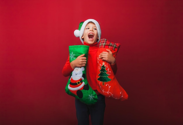 빨간 니트 스웨터와 산타클로스 모자를 쓴 소년이 선물용 크리스마스 부츠를 들고 있다 귀여운 아기가 크리스마스를 기다리고 있고 빨간 배경에 새로운 종류의 단열재가 있습니다