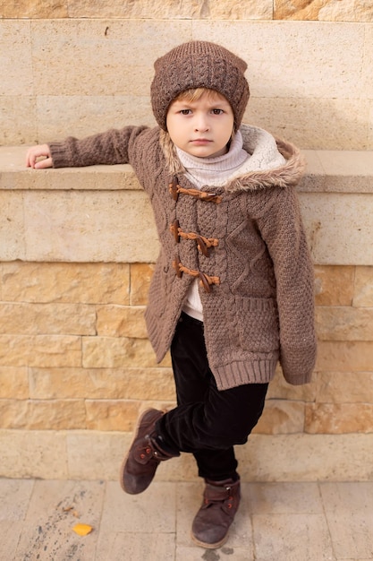 a boy in knitted things a hat and a brown jacket is standing on the street leaning against the wall