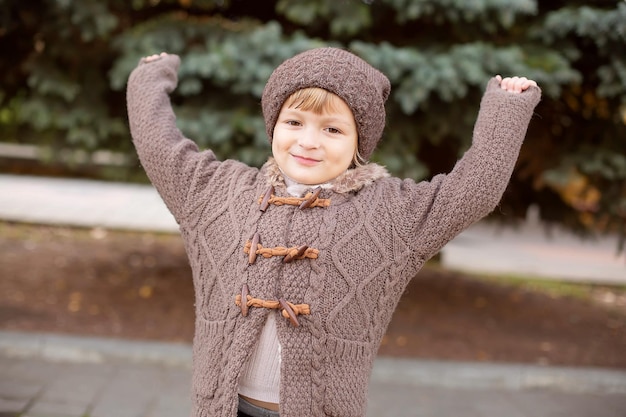 a boy in knitted things a hat and a brown jacket on the street against the background of a fir tree