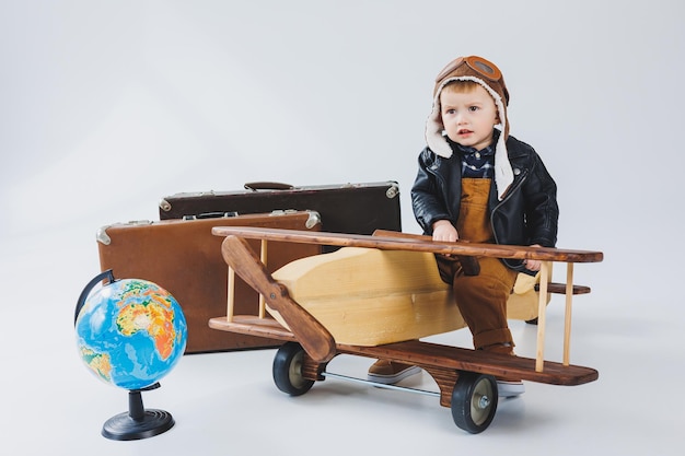 A boy in a leather jacket and a pilot's hat a wooden plane a globe brown suitcases Children's wooden toys