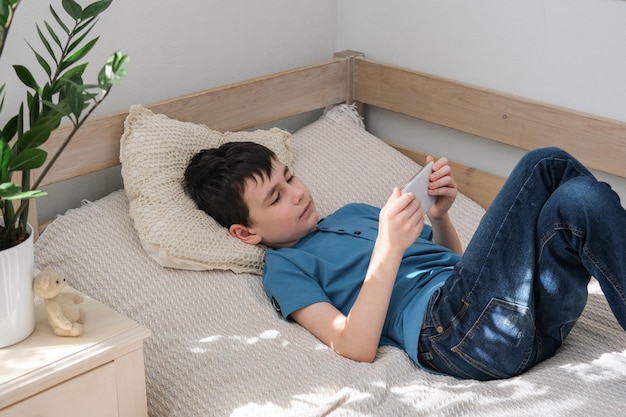 The boy lies on the bed and watches a movie or video on a smartphone Technology leisure for children