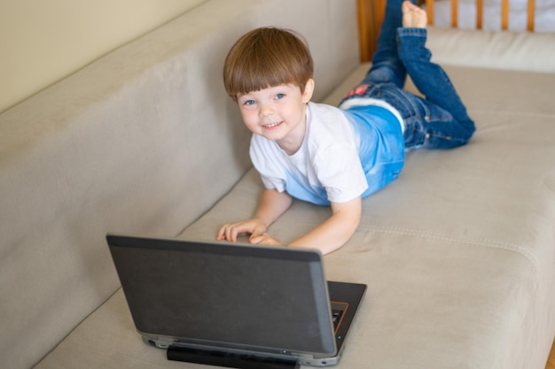 The boy lies on a bright sofa and looks at a laptop.