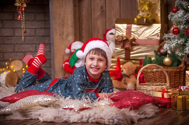 The boy lies on the floor near the Christmas tree among the boxes with Christmas gifts