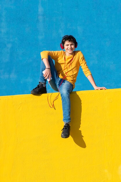 Boy listening to music through headphones while sitting on yellow railing against blue wall