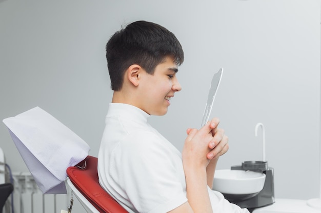 Boy looks in the mirror with a toothy smile sitting on a chair with a dentist in the dental office