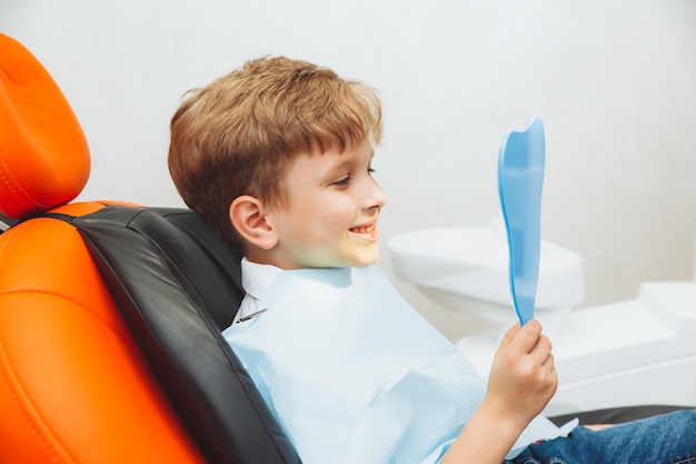 Boy looks in the mirror with a toothy smile sitting on a chair with a dentist in the dental office