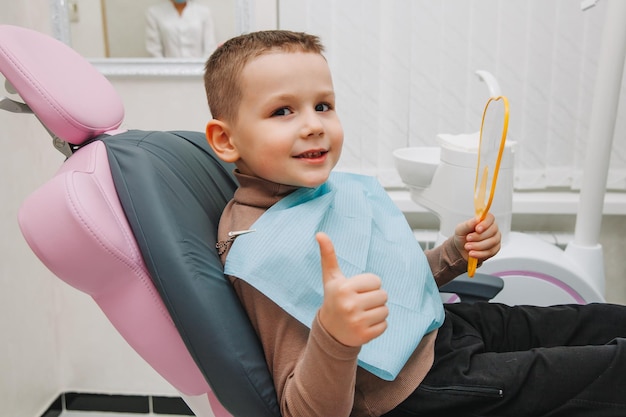 Boy looks in the mirror with a toothy smile, sitting on a chair with a dentist in the dental office.