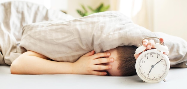 Boy lying on the bed under a blanket and stopping alarm clock in the morning