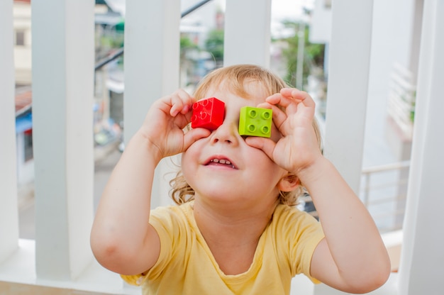 男の子はカラフルな子供のブロックの目を作ります。屋内でカラフルなプラスチック製のブロックをたくさん遊んでメガネでかわいい子供男の子。スキルと創造性の促進