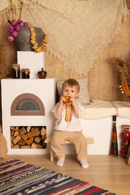 A boy in a national costume is sitting near a Russian stove and eating a bagel Pancake day