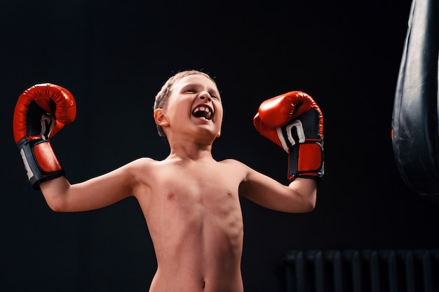 Un ragazzo vicino a un sacco da boxe in guantoni da boxe urla e alza le mani come un vincitore di latta.