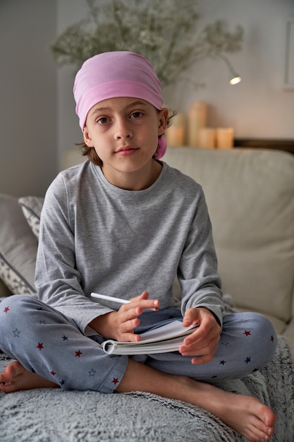 Photo boy in nightwear and pink headscarf for raising awareness of breast cancer sitting with crossed legs and writing in notepad while looking at camera