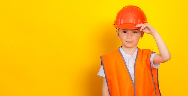 A boy in an orange construction helmet and a reflective vest stands against a yellow background