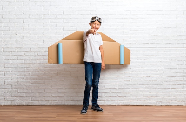 Boy playing with cardboard airplane wings on his back points finger at you