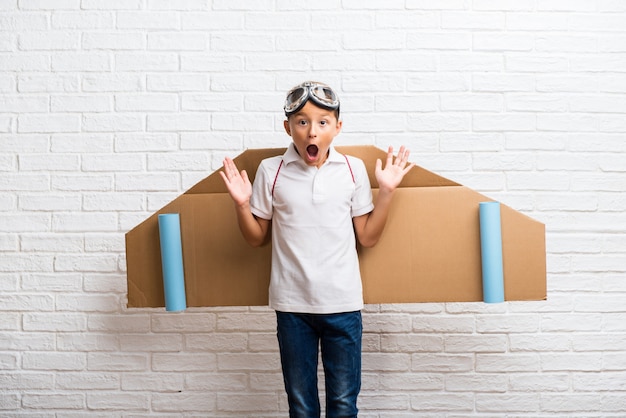 Boy playing with cardboard airplane wings on his back with surprise and shocked facial expression