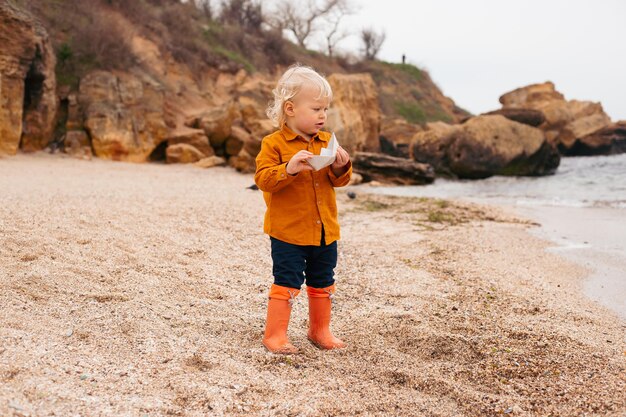 Boy playing with paper boat on the beach near sea in autumn or summer time