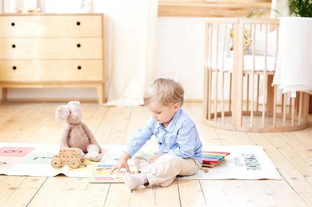 The boy plays with wooden toys at home. Educational wooden toys for the child. Portrait of a boy sitting on the floor in the children's room in the Scandinavian style. Eco toys, children's room decor