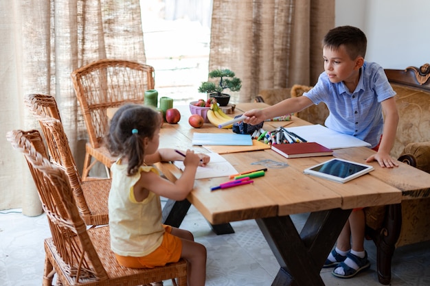 The boy, a primary school student, does his homework with his younger sister, helps her draw, and hands out a blue felt-tip pen.