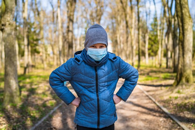 保護マスクをした少年が公園の小道に立っている子供が両手を腰に当てた10代の若者がパンデミックの状況に不満を持っている