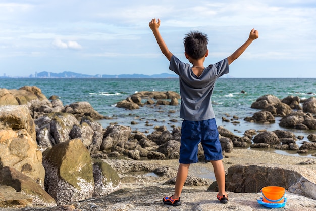 Boy raises his hands while standing on top rock looking at the sea.