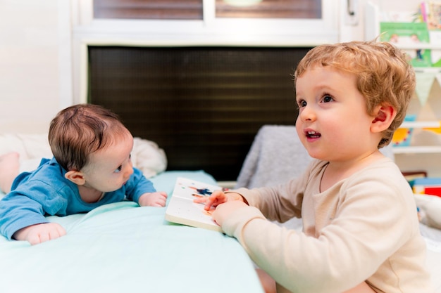 Boy reading book by sibling on bed at home