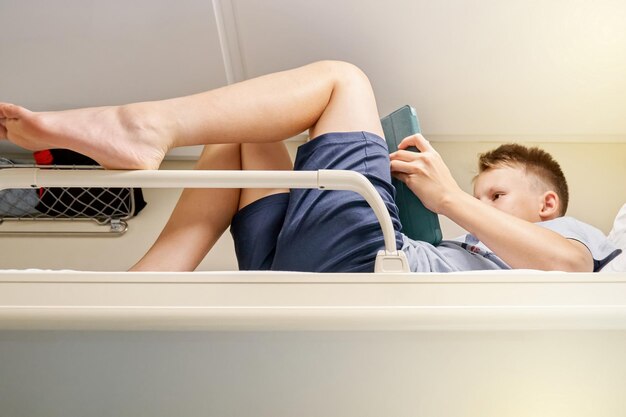 Boy reads electronic book via tablet PC lying on bedding on train car upper shelf low angle shot