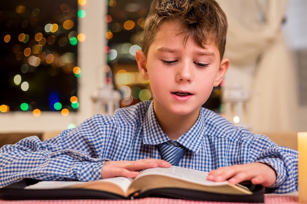 Boy reads a thick book. Child reading beside window. Reading is a good hobby. Interesting book of novels.