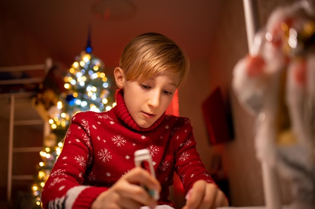 A boy in a red Christmas sweater on the background of a Christmas tree in his room finishes drawing for the new year