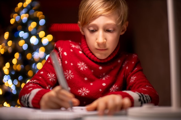 A boy in a red Christmas sweater on the background of a Christmas tree holds a marker in his hand and draws on paper