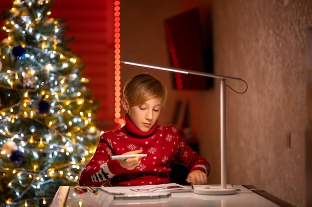 A boy in a red Christmas sweater on the background of a Christmas tree illuminated by a lamp holds a drawing marker in his hand