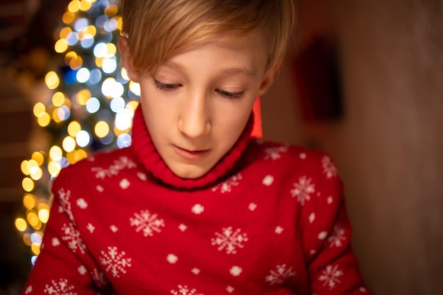 A boy in a red christmas sweater on the background of a\
christmas tree is engaged in the process of drawing in his room\
under a lamp