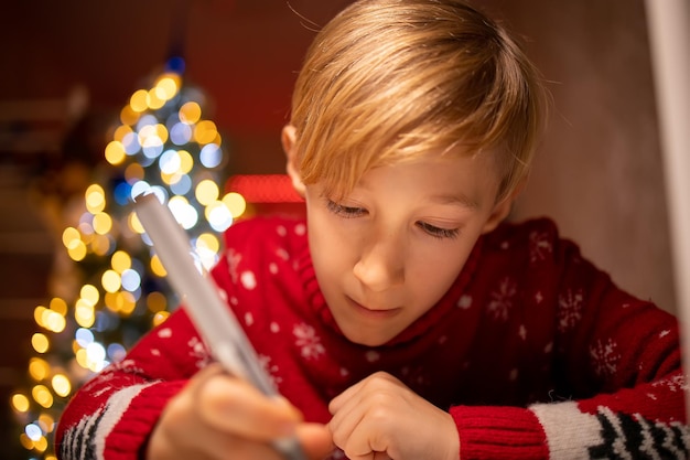 A boy in a red Christmas sweater on the background of a Christmas tree leans close to the table and tries to draw a drawing