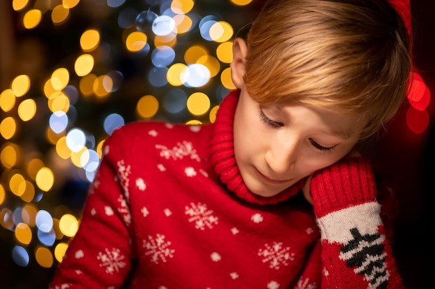 A boy in a red christmas sweater on the background of a\
christmas tree pressed his hand to his face and thoughtfully\
draws