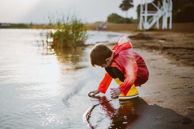 Мальчик в красном плаще и желтых резиновых сапогах играет с водой на пляже Школьник в непромокаемой куртке трогает воду в море Ребенок веселится с волнами на берегу
